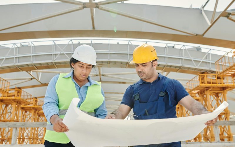 Male and female engineers discussing blueprints at a construction site, promoting teamwork and planning.